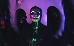 A group of people in black masks standing in front of an illuminated purple light. A group of people in black masks standing in front of an illuminated purple light.