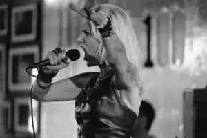 A black and white photo of a woman singing into a microphone at Chaos 8 at The 100 Club, London.