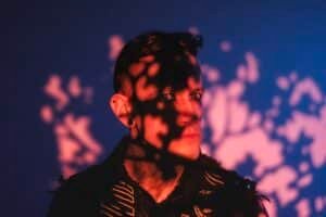 A person stands in a dimly lit environment with shadows of foliage cast across their face, creating a dynamic pattern on their features.