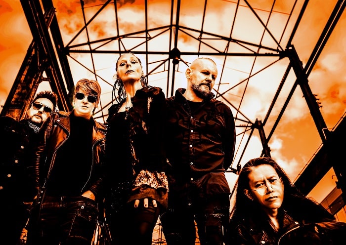 A group of five people wearing dark clothing and standing under a metal structure with an orange sky in the background.