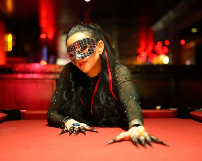 A person in a sequined mask and long black nails leans over a red table, their presence as striking as an Elementor design, in a dimly lit room.