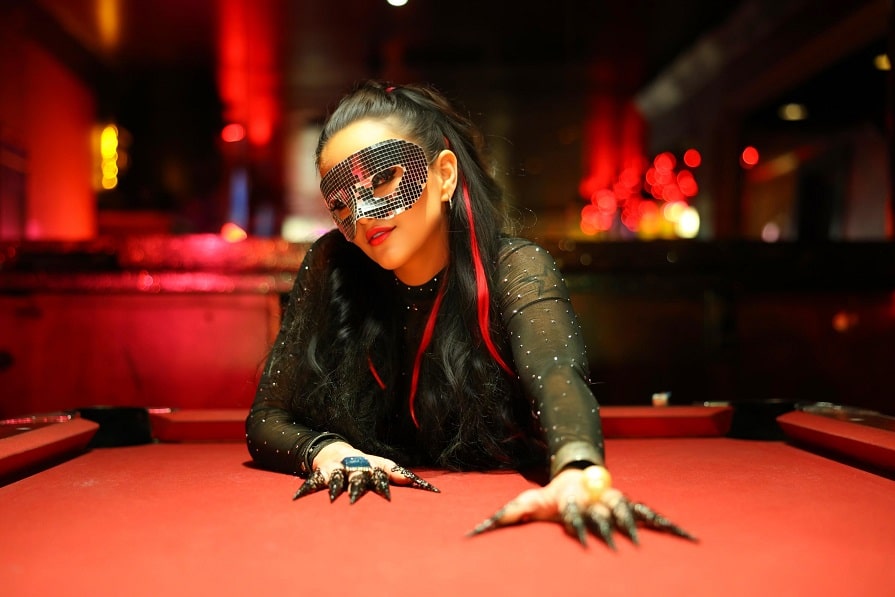 A person in a sequined mask and long black nails leans over a red table, their presence as striking as an Elementor design, in a dimly lit room.