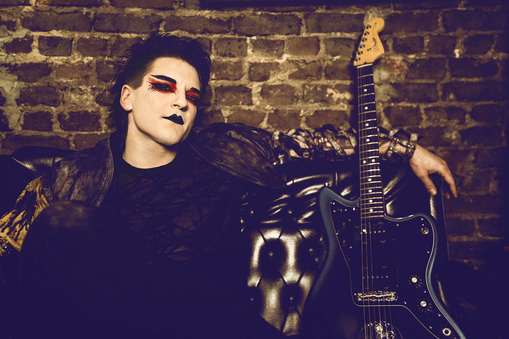 A person with bold makeup and a guitar, embodying the edgy style of Vision Video, leans against a brick wall on a leather couch.