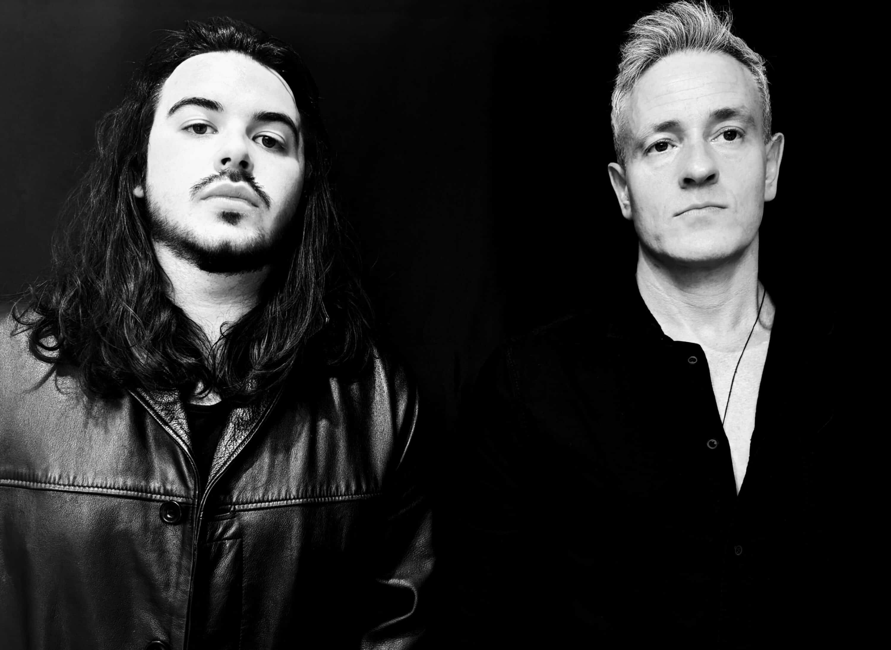 In a striking black and white photo, one man with long hair and a leather jacket stands beside another with short hair in a collared shirt. Their distinct styles create an elementor of contrast against the dark background.