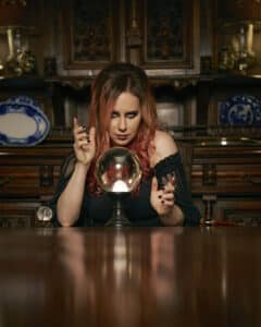 A woman with pink hair sits at a wooden table, focusing on a crystal ball, channeling her inner Miss FD in a room adorned with ornate furniture and decorative plates. Perhaps she’s crafting the next release—a spellbound review of a curse breaker.