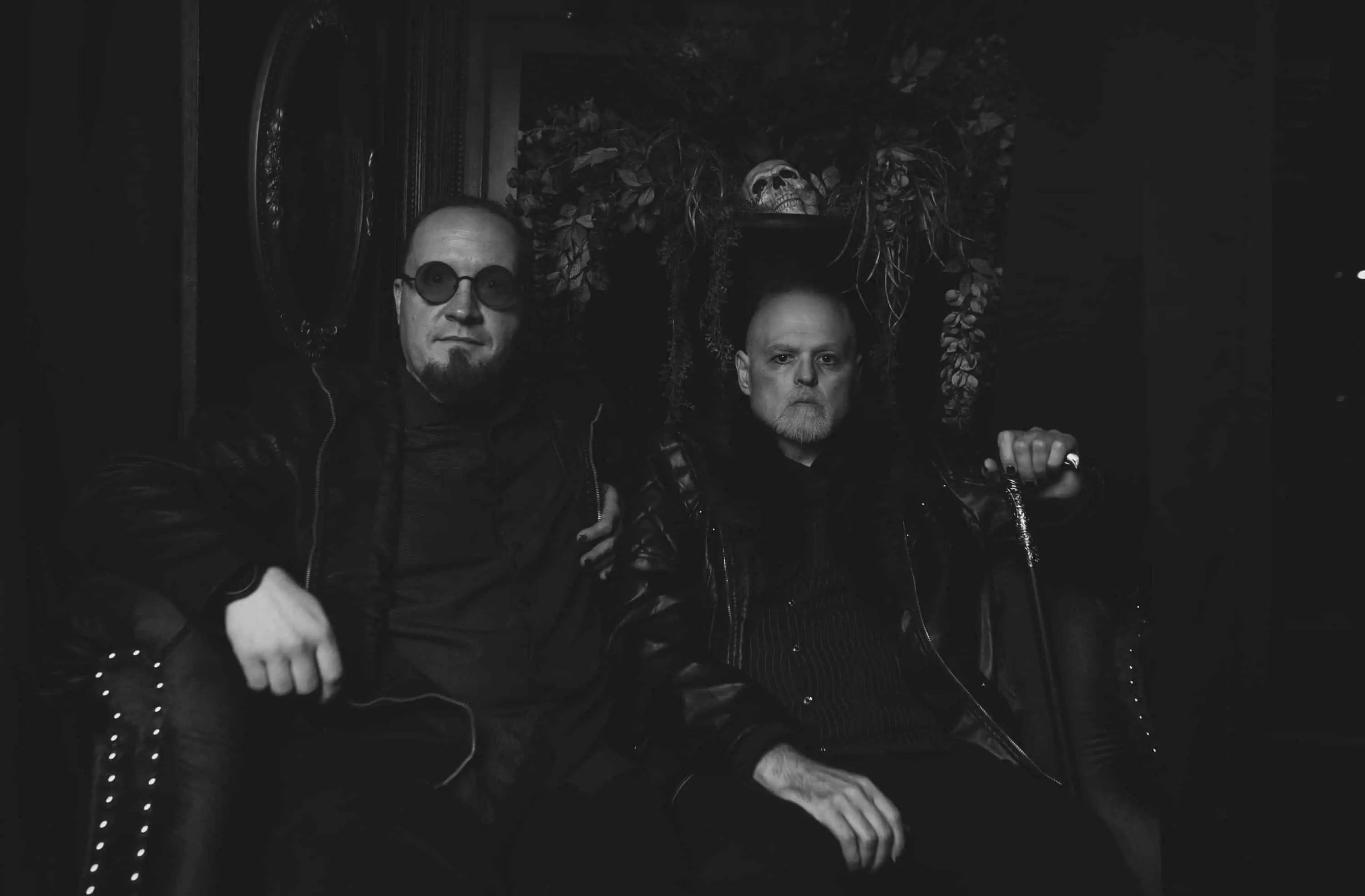 Two men in dark clothing sit close together in a dimly lit room, exuding a vibe that screams "It All Falls Apart." Both wear glasses and leather jackets; one grips a cane, embodying the essence of a Funeral March. A decorative display looms hauntingly in the background.