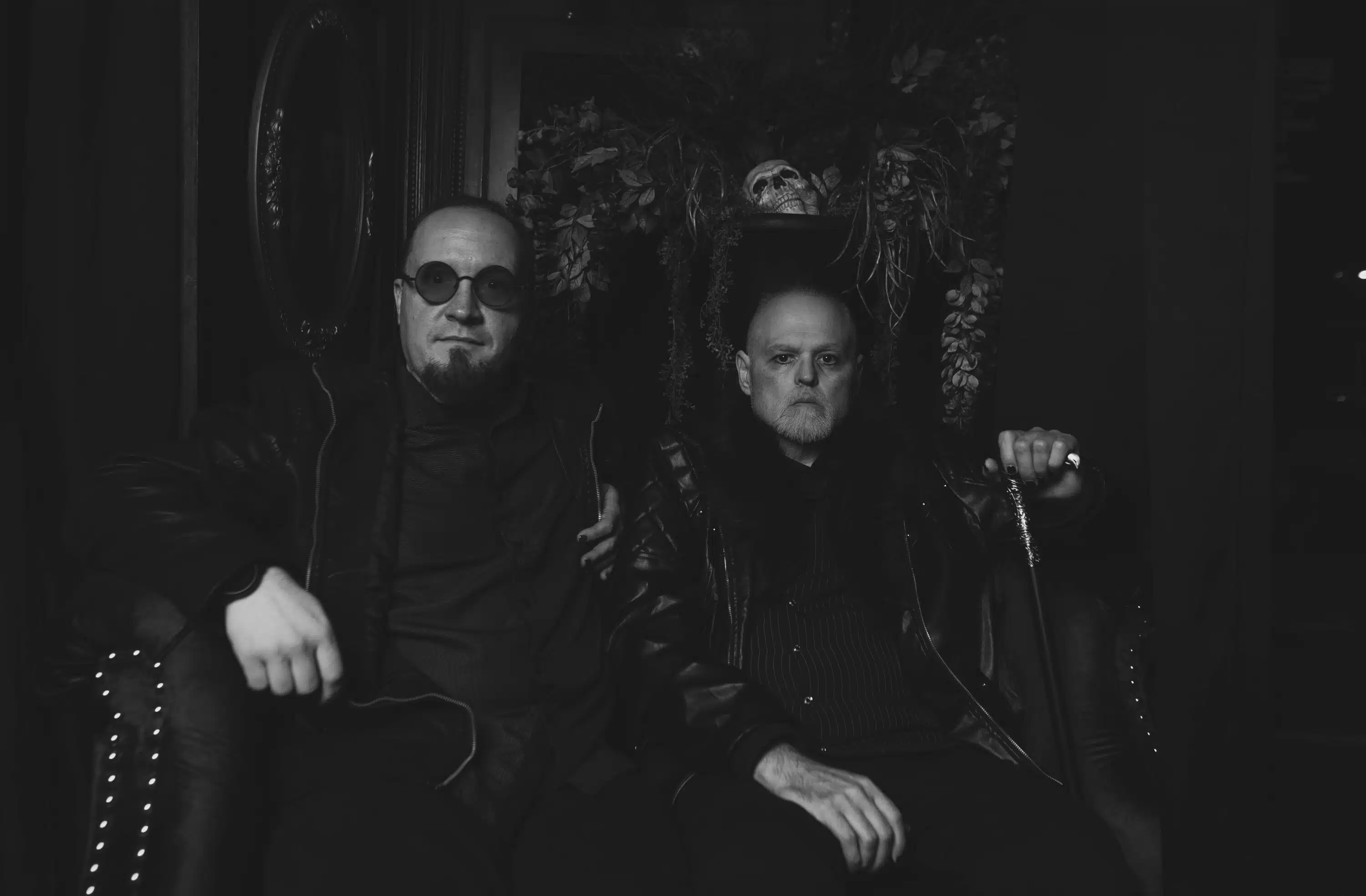 Two men in dark attire sit in a dimly lit room. One wears sunglasses, and they both appear serious. Behind them is a floral arrangement with a skull, evoking themes from "The Funeral March" album release.
