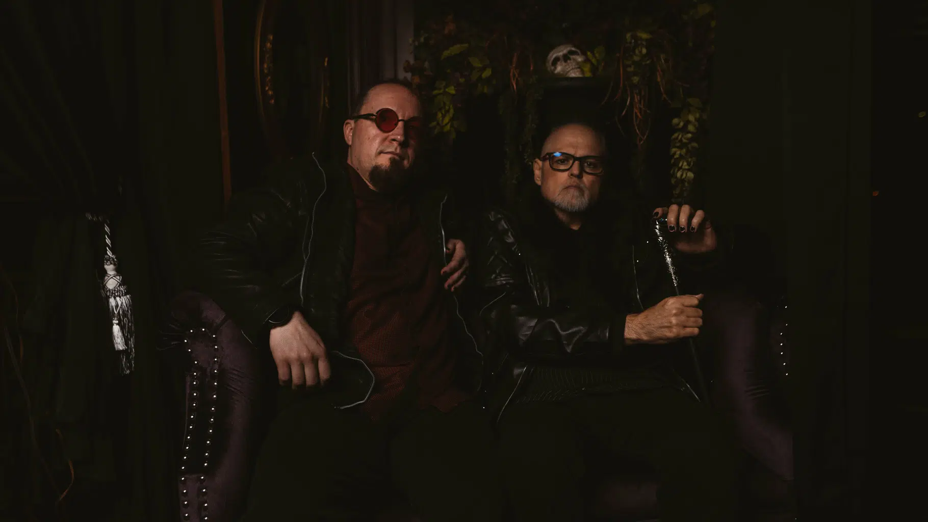 Two men in leather jackets and sunglasses sit on a purple couch, one holding a cane. The dimly lit backdrop with foliage and a skull adds a touch of drama, reminiscent of an "It All Falls Apart" album cover vibe.
