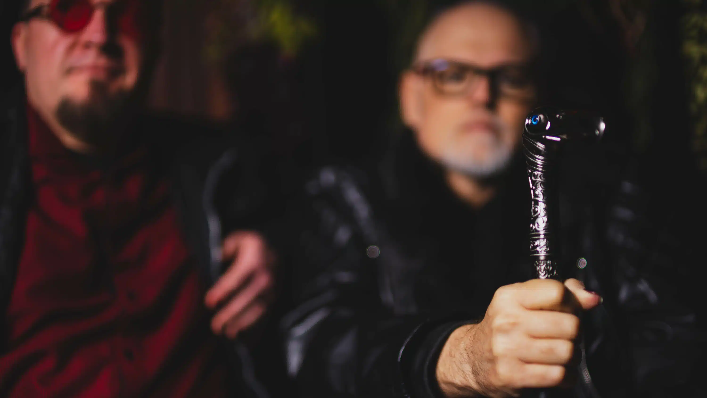 Two men in dark clothing, one on the left partially seen, and the other holding an intricately designed cane towards the camera. Their pose mirrors the contemplative mood of a Funeral March album release review, as they sit closely together.
