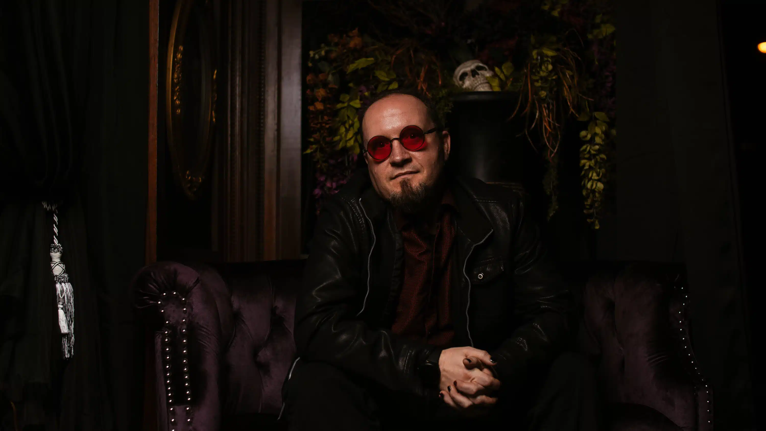 A man in red sunglasses and a leather jacket lounges on a purple couch, evoking the mood of a "Funeral March" album review, while plants and a skull cast shadows in the dimly lit room.