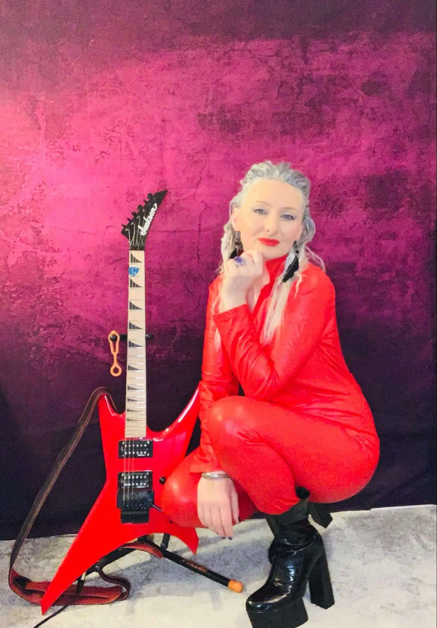 Person in a red outfit and black boots crouches beside a red electric guitar against a purple background, ready for their interview. The scene is reminiscent of "The Eyes Of Heisenberg," with its vivid hues and captivating presence.