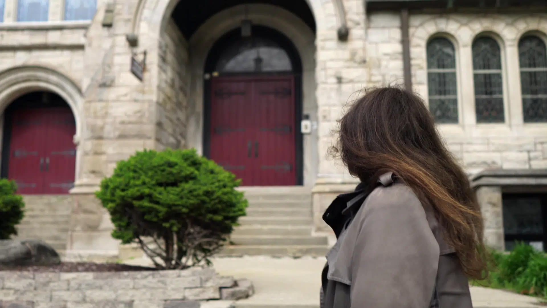 A person with long hair in a coat stands before a stone building with a red arched door, contemplating it as if pondering the somber notes of a Funeral March. To the left, a small bush adds a touch of life to the scene.