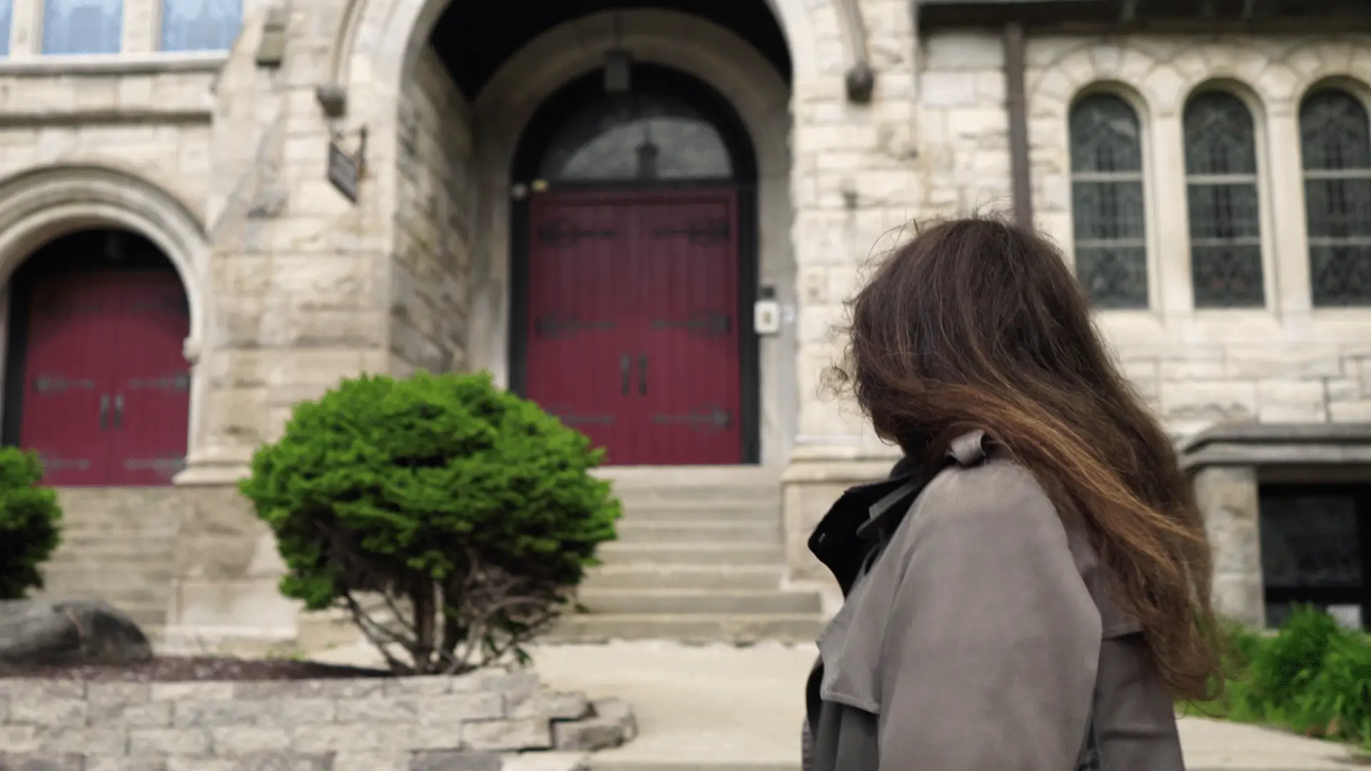 A person with long hair in a coat stands before a stone building with a red arched door, contemplating it as if pondering the somber notes of a Funeral March. To the left, a small bush adds a touch of life to the scene.