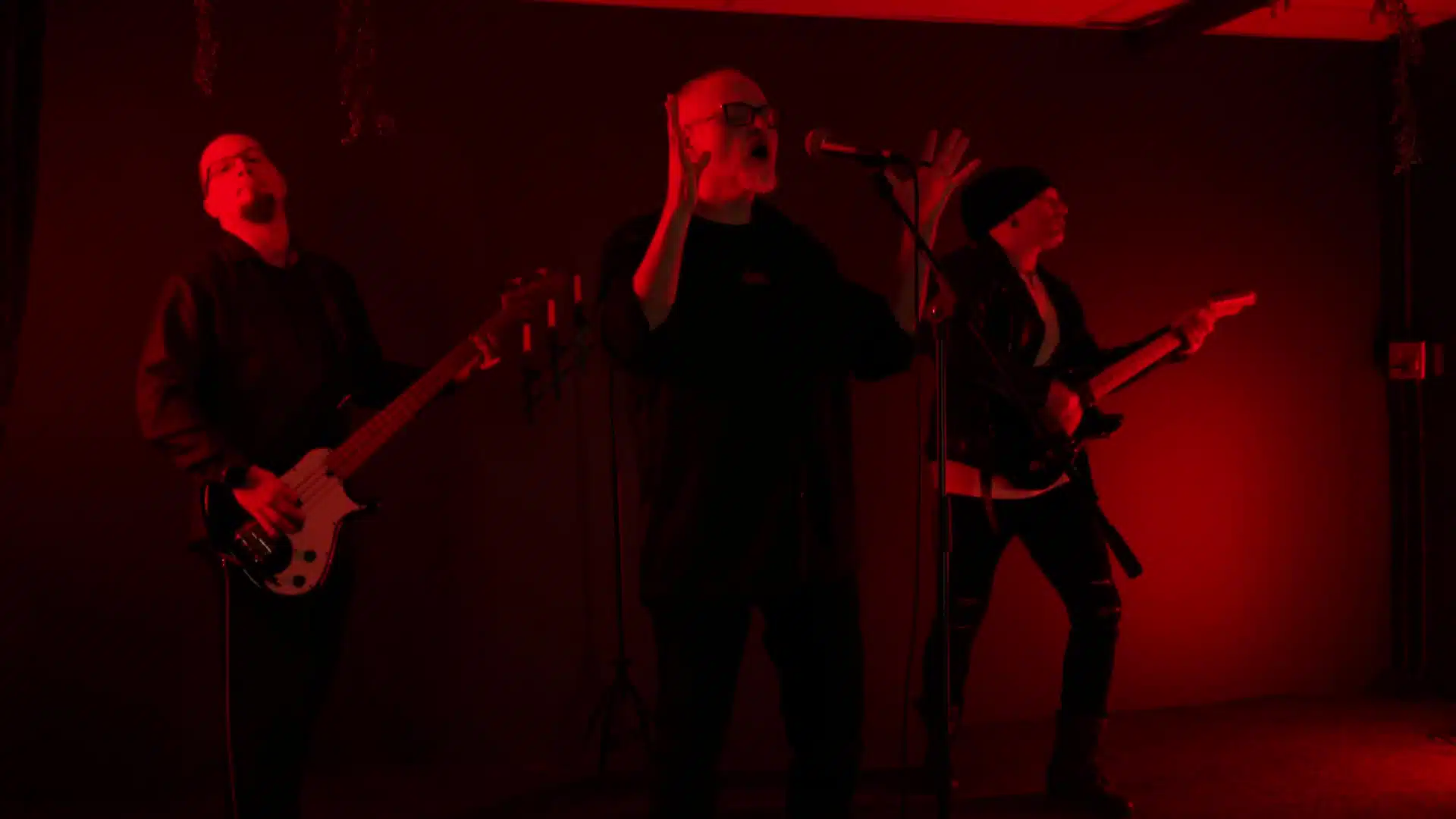 Three musicians perform on stage under red lighting, reminiscent of a somber Funeral March. A singer stands center stage, flanked by a bassist on the left and a guitarist on the right, creating an atmosphere where it all falls apart beautifully.