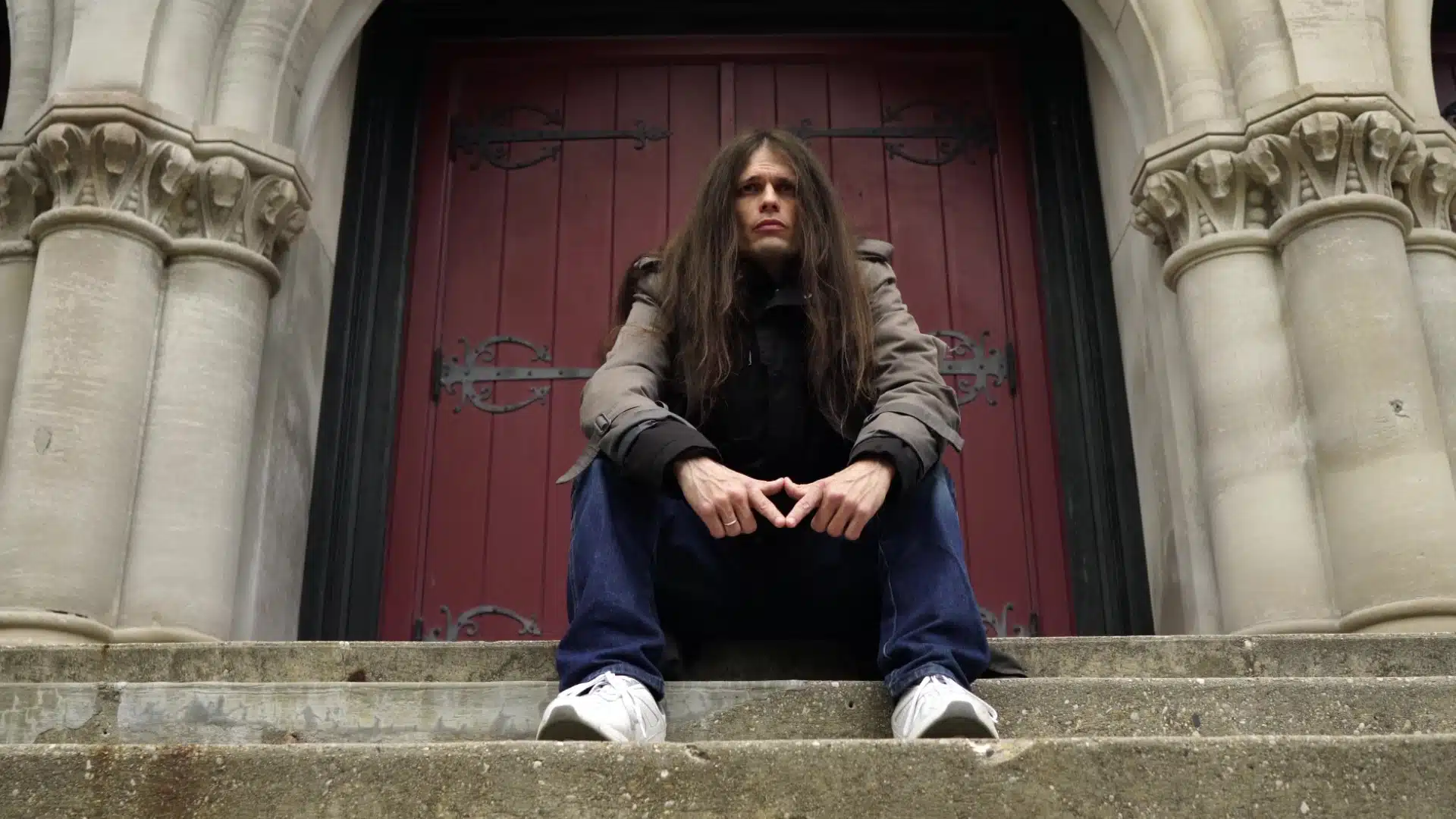 Person with long hair seated on stone steps in front of a large red door with ornate hinges, wearing a gray jacket, jeans, and white sneakers, as if pondering an "It All Falls Apart" album review.