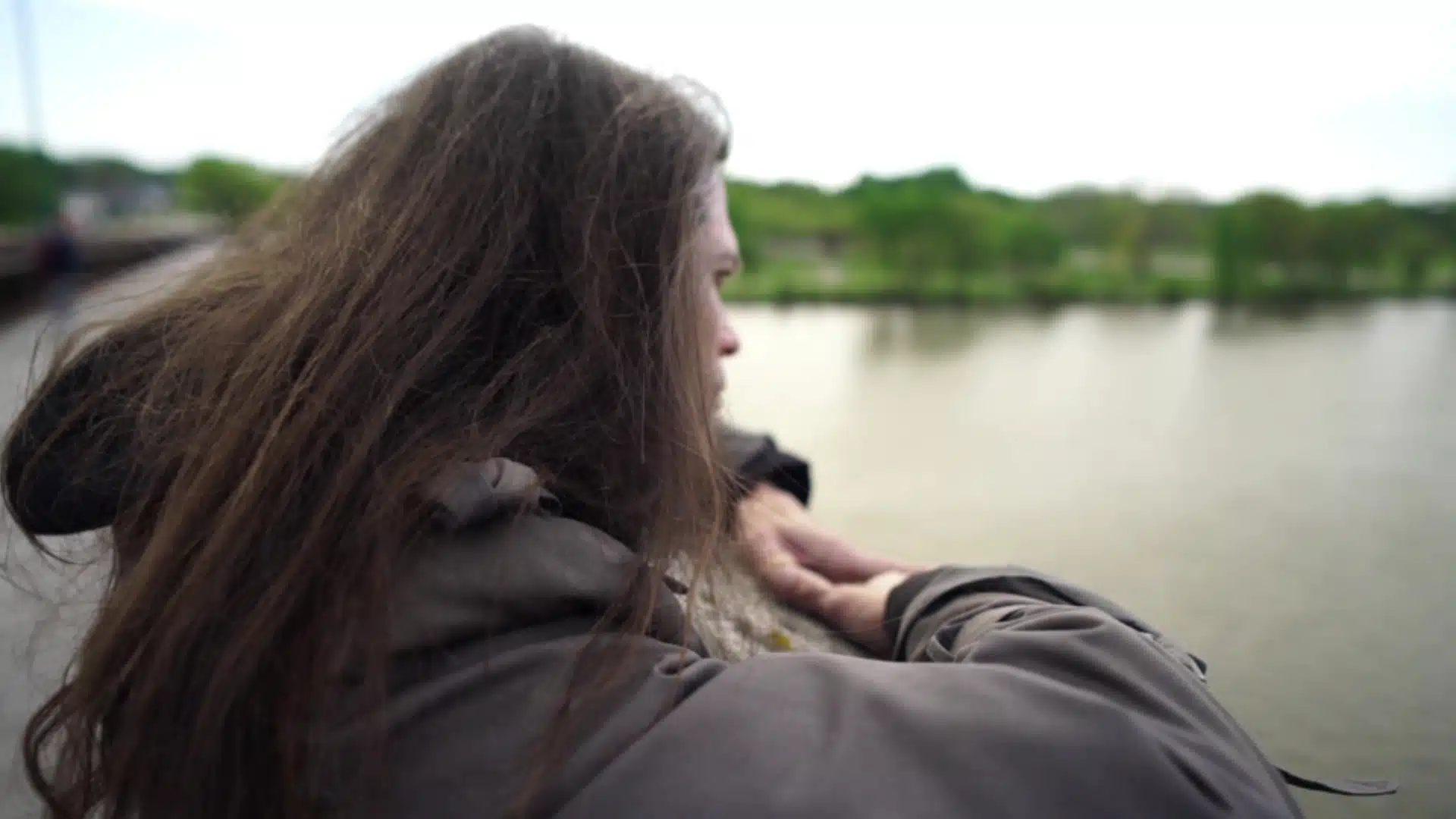 Person with long hair and a jacket stands by a railing, contemplating like a mournful Funeral March, as they gaze over the water with trees in the background.
