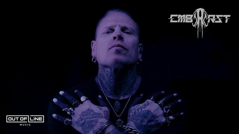A tattooed man with piercings and multiple rings crosses his arms over his chest against a dark background, with "Combichrist" and "Out of Line Music" logos visible.