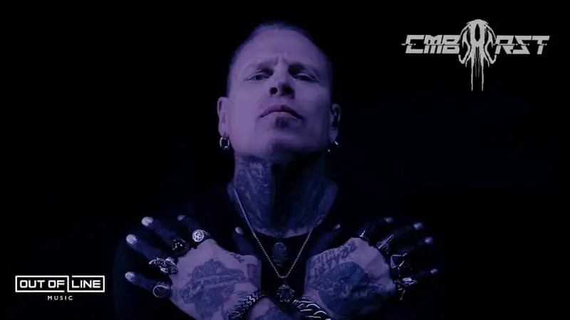 A tattooed man with piercings and multiple rings crosses his arms over his chest against a dark background, with "Combichrist" and "Out of Line Music" logos visible.