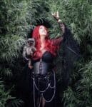 A woman with long red hair, dressed in black gothic attire with chains, stands among tall green foliage, holding a small dog and reaching up with one hand—like a cover from the Haunted Hearts XIII album.