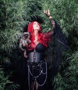A woman with long red hair, dressed in black gothic attire with chains, stands among tall green foliage, holding a small dog and reaching up with one hand—like a cover from the Haunted Hearts XIII album.