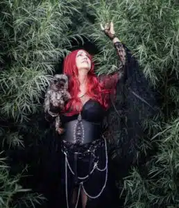A woman with long red hair, dressed in black gothic attire with chains, stands among tall green foliage, holding a small dog and reaching up with one hand—like a cover from the Haunted Hearts XIII album.