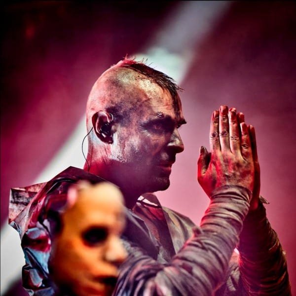 A person with a shaved head, face paint, and dirty hands clasps their hands on stage as FIX8:SED8 performs; another masked figure stands in the foreground, dramatic red lighting hinting at their Octagram album's intense themes.