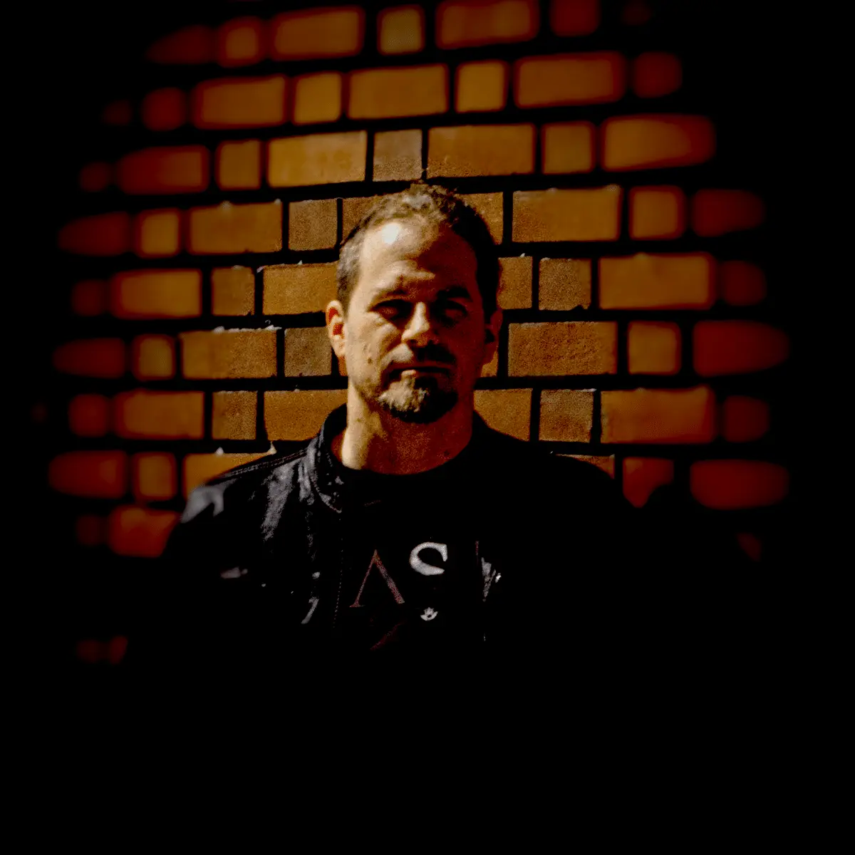 A man with short hair and a beard stands in front of a brick wall, wearing a black jacket. The lighting is dim and focused on his face and upper body.