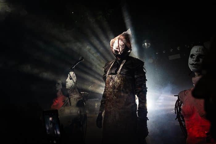 Three people in unsettling costumes and masks stand on a dimly-lit stage, with dramatic rays of light shining behind the central figure.