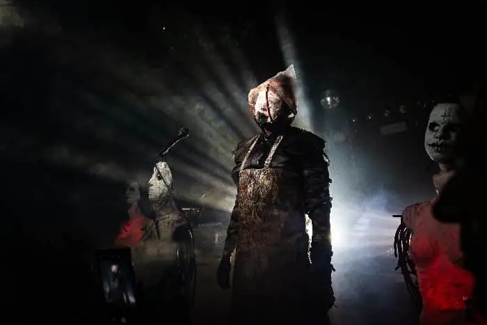 Three people in unsettling costumes and masks stand on a dimly-lit stage, with dramatic rays of light shining behind the central figure.