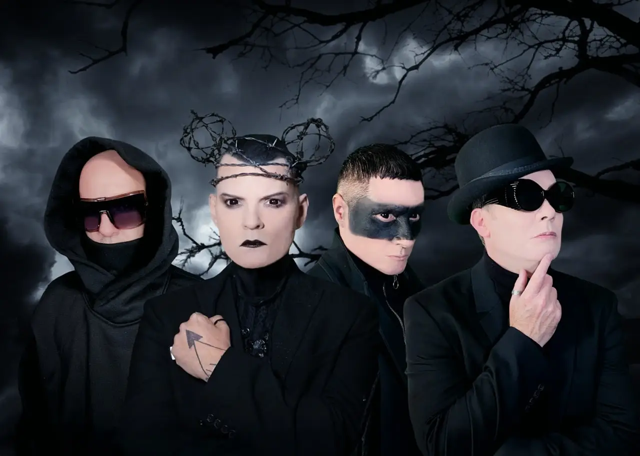 Four people dressed in black with dramatic make-up and accessories stand in front of a dark, cloudy sky with bare tree branches.