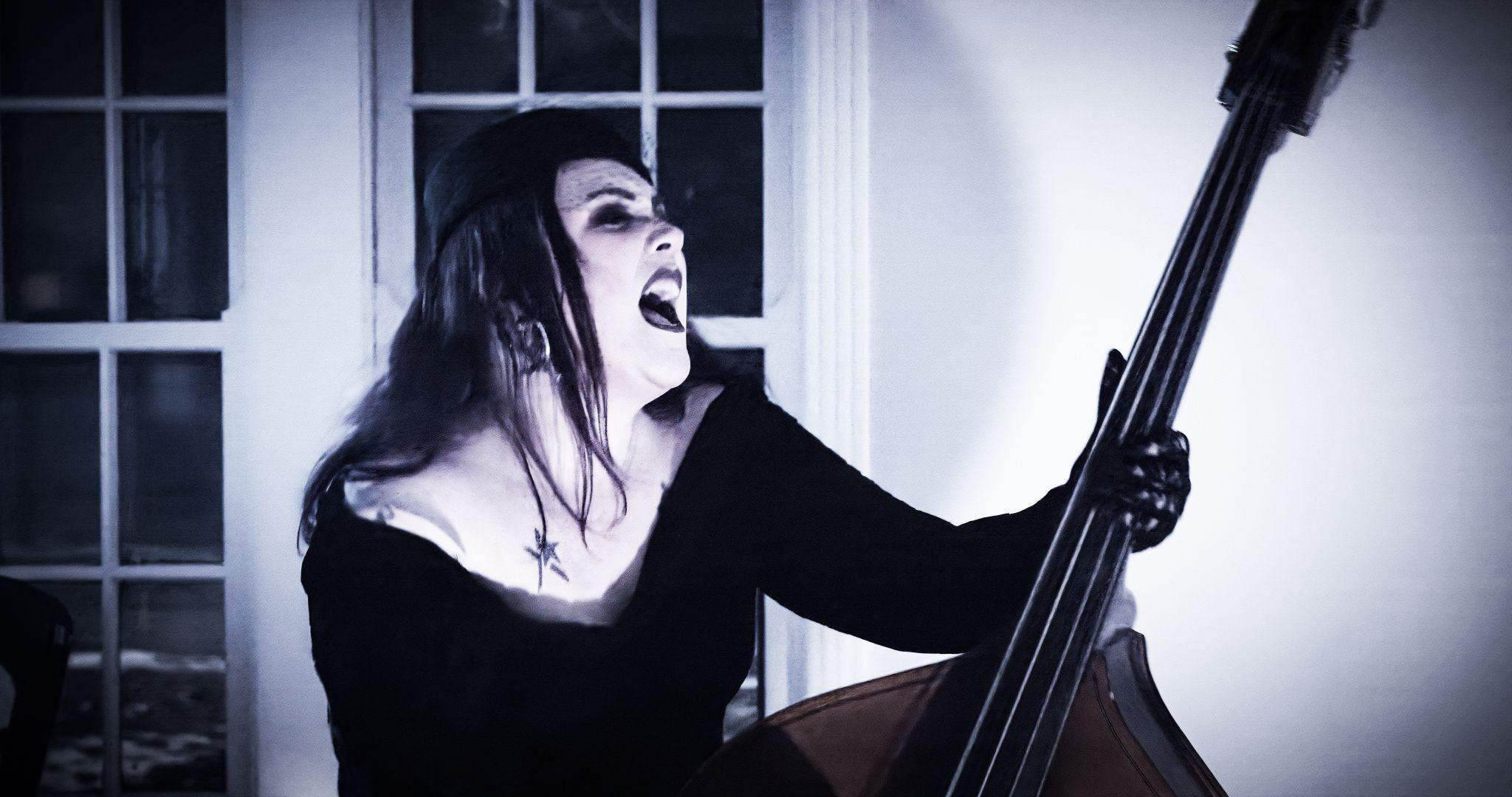Person wearing dark clothing and gloves plays an upright bass passionately indoors near large windows.