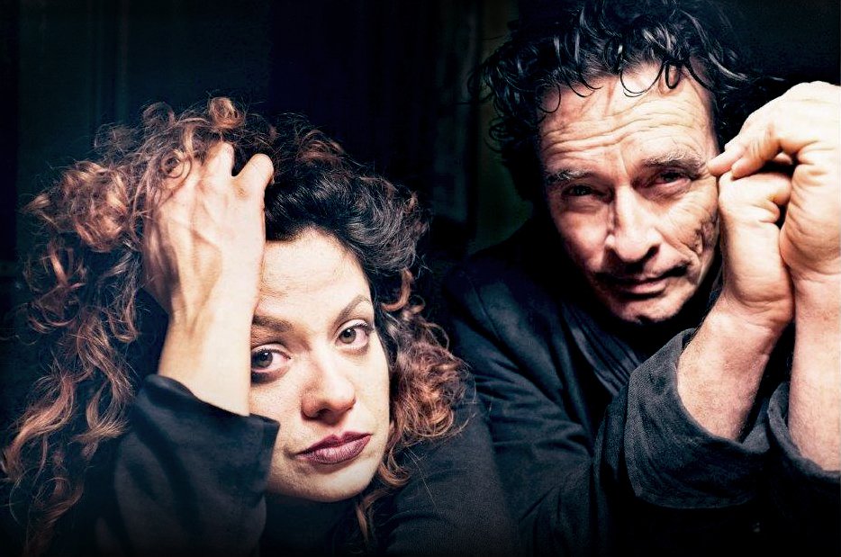A woman and a man with curly hair sit close together indoors, both looking towards the camera with neutral expressions. The lighting is dim and dramatic.