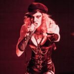 A performer in a black leather outfit, gloves, and cap sings into a microphone under red stage lighting, with one hand reaching forward—a striking visual from Numa Echos' "Descending Consciousness" album review.